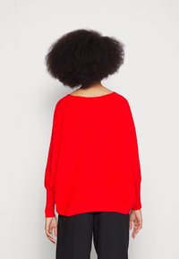 Pull en tricot rouge à coupe ample, avec un col large et des manches longues. Le matériau semble doux avec une texture côtelée, se terminant au niveau des hanches.