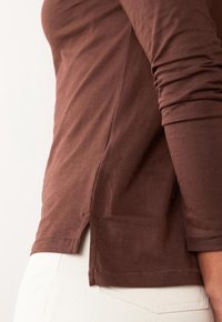 Brown long-sleeve shirt made of soft fabric, features a side slit at the hem and fitted sleeves, paired with white pants.