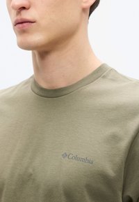 Πράσινο ελαιόχρωμο βαμβακερό μπλουζάκι με στρογγυλή λαιμόκοψη και κοντά μανίκια, με διακριτικό λογότυπο Columbia τυπωμένο σε μπλε στο στήθος.