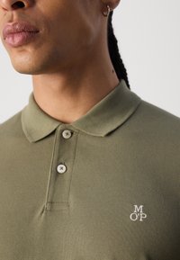 Cămașă polo de culoare verde măslinie cu un material texturat, cu trei nasturi și un mic logo brodat pe pieptul stâng.