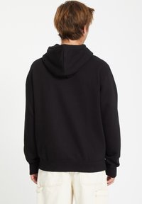 Zwarte hoodie van zacht materiaal, met een capuchon en geribbelde details bij de manchetten en de zoom, gedragen over lichtgekleurde cargobroeken.