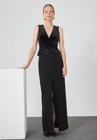 Top sin mangas de terciopelo negro con escote en V combinado con una falda negra de alta abertura. Modelo con tacones negros de pie junto a un pedestal blanco.