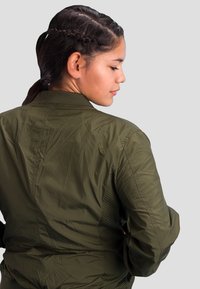 Olive green lightweight bomber jacket with textured side panels, ribbed collar, and visible seams; fitted design and functional style.