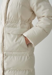 Manteau doudoune beige avec une texture lisse, des coutures horizontales et des poches latérales ; présente une taille cintrée et de larges poignets.
