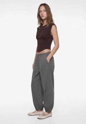 Giovane donna che indossa un top marrone senza maniche, pantaloni larghi a quadri con polsini elastici e scarpe basse beige, in piedi con una mano in tasca.