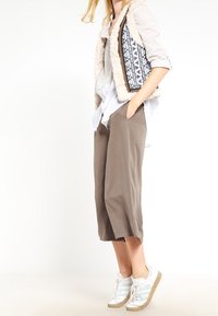 Gilet in pelliccia sintetica beige con rivestimento blu a fantasia, camicia bianca, culotte grigie e sneaker chiare con dettagli beige.