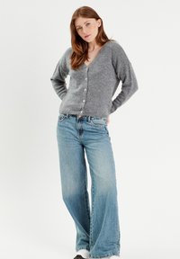 Cardigan grigio a maglia con scollo a V, caratterizzato da maniche lunghe e sei bottoni frontali. Indossato con jeans a gamba ampia di colore blu chiaro.