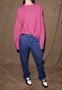 Pull rose surdimensionné, jeans bleu à jambes droites et chaussures noires et blanches. Texture douce, coupe décontractée et design casual, sur un fond marron.