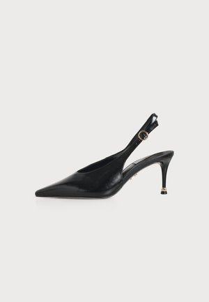 Escarpin slingback en cuir noir à bout pointu, talon fin et bride de cheville ajustable avec boucle ronde dorée sur fond blanc.