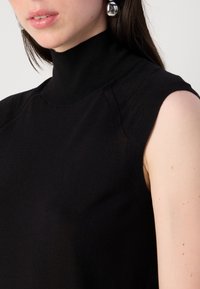 Detailní záběr ženy, která má na sobě černý bezramínkový top s vysokým výstřihem, a v uchu jsou viditelné stříbrné kulaté náušnice.