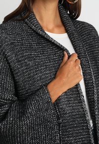 Eine Frau trägt eine strukturierte, dunkelgraue Strickjacke über einem weißen Shirt, hält den Kragen mit einer Hand und zeigt Creolen.
