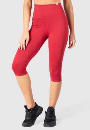 Czerwone legginsy capri wykonane z gładkiego, elastycznego materiału z wysokim stanem, w połączeniu z czarnymi butami sportowymi. Szczegóły szwów na nogawkach.