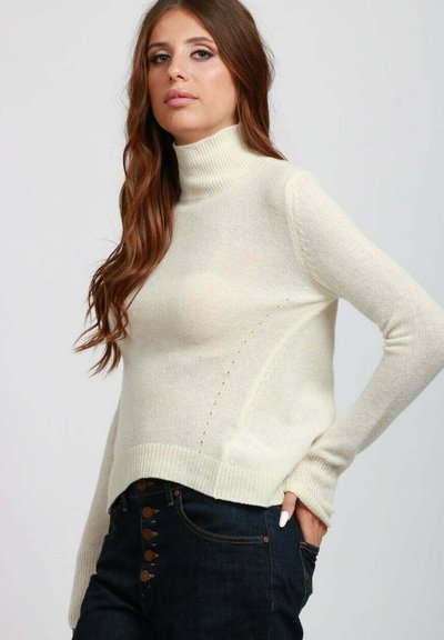 Donna con lunghi capelli castani che indossa un maglione a collo alto color crema e jeans blu scuro con dettagli ricamati, in posa con una mano in tasca.