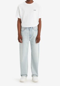 T-shirt bianca in cotone con logo nero, abbinata a jeans in denim azzurro chiaro. Il modello indossa sneakers bianche. Design semplice; vestibilità rilassata.