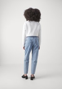 Persona con cabello rizado y voluminoso, vistiendo un sudadera blanca, jeans de mezclilla claros y zapatos de tacón negros, de pie de espaldas sobre un fondo blanco.