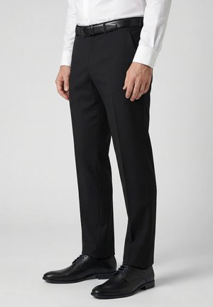 Mand iført sorte habitbukser, sort bælte, hvid skjorte og sorte pæne sko stående foran en ensfarvet baggrund.