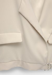 Bež blazer izrađen od glatke tkanine, s rebrastim ovratnikom, dva prednja džepa i detaljem u obliku srebrnog metalnog prstena u struku.