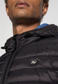 Dolna część twarzy i szyja osoby noszącej czarną pikowaną kurtkę z zamkiem i naszywką z logo w pobliżu ramienia, widoczny kaptur wyściełany niebieską tkaniną.