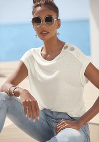 Witte short-sleeve top met een comfortabele pasvorm, voorzien van drie decoratieve knopen op de schouder. Gekleed met lichtblauwe jeans, tegen een kustdecor.