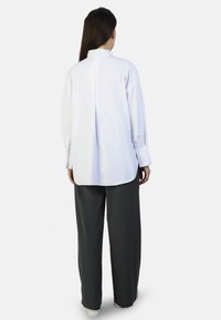 Long-sleeve white shirt with a back pleat, paired with loose-fitting dark gray trousers. Smooth fabric with buttoned cuffs and a rounded hem.