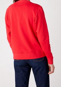Sweatshirt rouge avec un col rond et des manches longues, présentant une texture lisse et une coupe décontractée, associé à un jean bleu foncé.