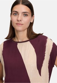 Junge Frau mit glattem braunem Haar, die ein lila-beige gemustertes Kurzarmoberteil vor einem schlichten Hintergrund trägt.