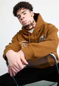 Sudadera marrón con un logo, detalles en texto blanco y cordón. Modelo sentado, con los brazos apoyados en las piernas, vistiendo pantalones negros y sentado en una silla transparente.