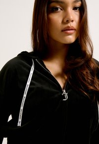 Hoodie preto com fecho de correr, apresentando um tecido texturizado, cordões brancos com marca e um detalhe em logo prateado no zíper.