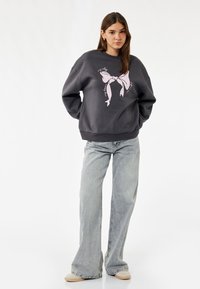 Grå sweatshirt med et pink sløjfe grafik på forsiden, der har en afslappet pasform, ribbede manchetter og rund hals. Sammen med lyseblå jeans.