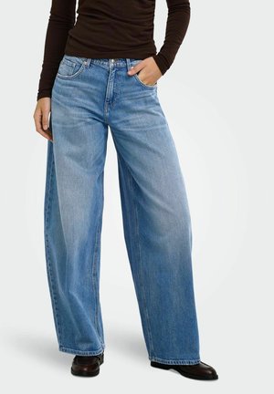 Person iført vidde blå jeans og sorte sko, med den ene hånd i lommen og en mørk langærmet top synlig på overkroppen.