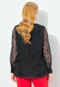 Blusa nera con maniche trasparenti a pois, tessuto testurizzato, scollatura rotonda e taglio dritto; presenta bottoni ai polsini e un orlo curvo.