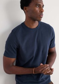 Tamsiai mėlynas medvilnės trumpa rankovė marškinėliai su apvalia iškirpte ir lygiu paviršiumi, dėvimi stovinčio asmens.