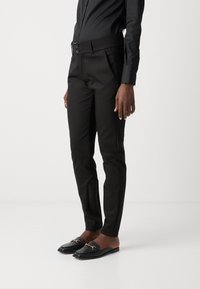 Pantaloni neri aderenti con una texture liscia, caratterizzati da una cintura alta e due bottoni. Abbinati a mocassini neri con dettagli in metallo dorato.