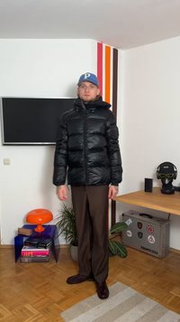 Casaco puffer preto com gola alta e fecho de zip, apresentando um acabamento brilhante. Combinado com calças castanhas e um boné de basebol azul.