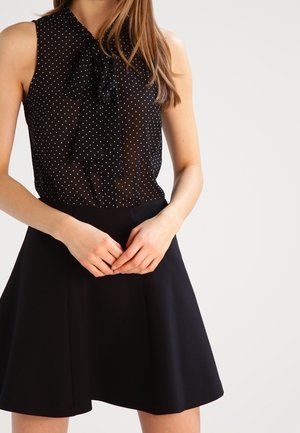 Blouse sans manches noire avec des pois blancs et un col à nœud papillon, associée à une jupe noire évasée. Tissu lisse avec une taille ajustée.