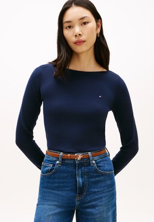 Kvinde med langt mørkt hår iført en mørkeblå langærmet top, brun bælte og blå jeans, stående mod en ensartet lys baggrund.