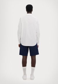 Camicia bianca a maniche lunghe con orlo arrotondato, abbinata a pantaloni corti blu scuro. Modello che indossa sneakers bianche e calzini grigi. Sfondo neutro.