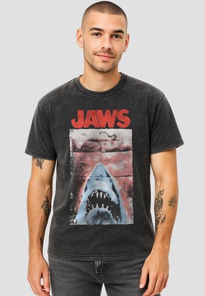 Young man with short hair and tattoos wears a black faded T-shirt with red "JAWS" text and a graphic of a shark approaching a swimmer.