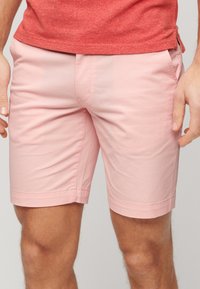 Hombre con pantalones cortos rosa claro hasta la rodilla y una camiseta corta de manga corta color rojo melange, de pie con las manos relajadas a los lados.