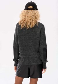 Grauer Strickpullover mit Lederelbow-Patches, geripptem Saum und langen Ärmeln. Getragen mit schwarzen Shorts aus Kunstleder.