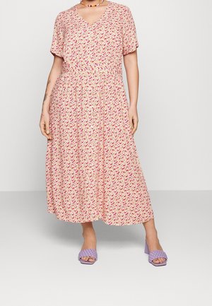 Kvinde iført en lys pink blomsterprint midi-kjole med korte ærmer, V-hals, perlehalskæde og lyserøde hælede sandaler.