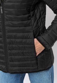 Svart vadderad jacka med horisontellt quiltat mönster, dragkedja framtill och sidofickor, kombinerad med blå jeans.