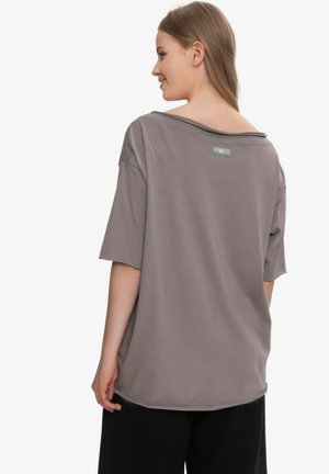 Kobieta z długimi włosami nosi luźny szary T-shirt i czarne spodnie, lekko obrócona bokiem, pokazując tył i szczegóły dekoltu T-shirtu.