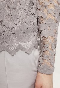 Grå blonde top med et blomsterprint, lange ærmer og scalloped kant, lagt over en lysegrå bluse.