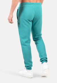Turkusowe spodnie dresowe o zwężanym kroju, z elastycznym pasem, kieszeniami po bokach oraz ściągaczami na nogawkach. Zestawione z białymi trampkami na neutralnym tle.