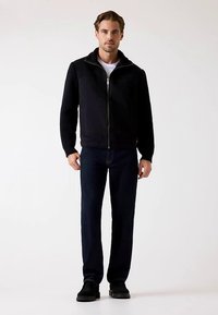 Črna jakna z zadrgo in rebrastimi rokavi, kombinirana s temno modrimi jeans hlačami in črnimi čevlji. Model pozira na nevtralni podlagi.