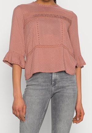 Lys pink bluse med en tekstureret stof, der har en firkantet yoke, indviklet blondekant og flagrende ærmer, kombineret med grå jeans.