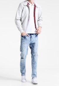 Mężczyzna w białej kurtce z czerwonym w kratkę podszewką, białej koszulce polo, jasnych dżinsach z przetarciami i białych sneakersach, stoi z jedną ręką w kieszeni.