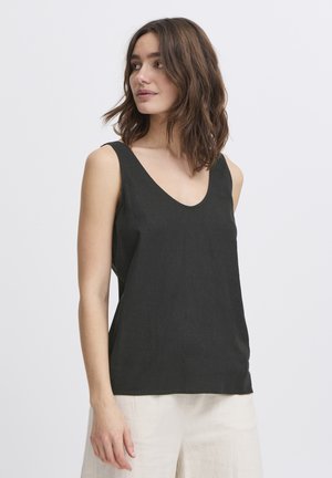 Femeie cu păr maro până la umeri, purtând un top negru fără mâneci și pantaloni deschiși la culoare, privind spre lateral pe un fundal simplu.