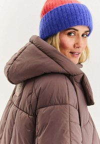 Brązowa pikowana kurtka puchowa z kapturem. Na głowie kolorowa dziergana czapka w różowym, niebieskim i fioletowym odcieniu.